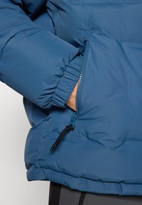Chaqueta puff azul con un bolsillo con cremallera y puños fruncidos. Textura suave y lisa con un acabado mate y acentos de hardware mínimos.