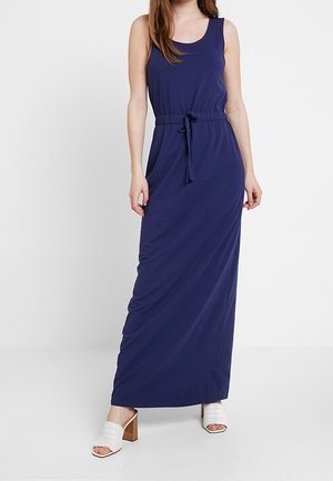 Robe maxi bleu marine sans manches avec ceinture à nouer à la taille, portée avec des sandales blanches à talons ouverts sur un fond uni.