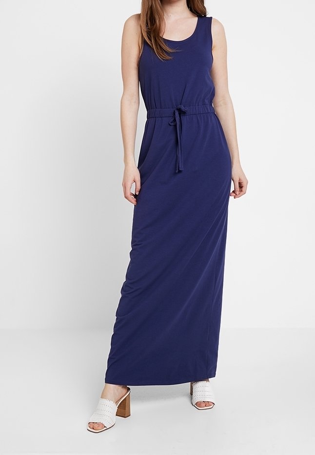 Robe maxi bleu marine sans manches avec ceinture à nouer à la taille, portée avec des sandales blanches à talons ouverts sur un fond uni.