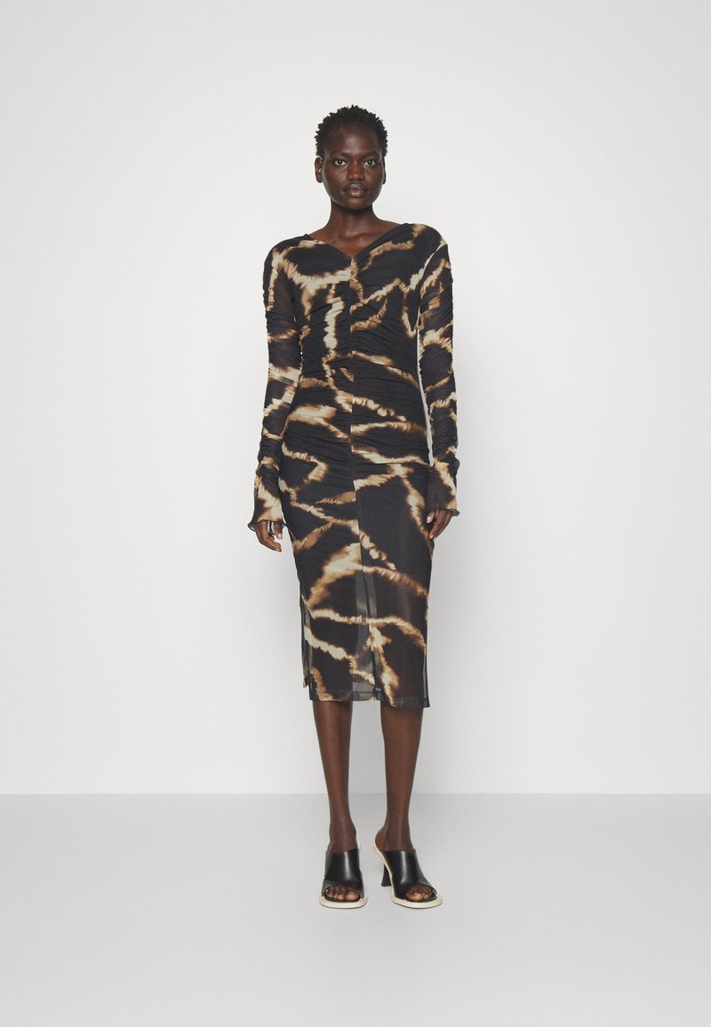 Robe tie-dye noire et marron à manches longues avec des détails froncés, forme ajustée et une fente sur le côté. Associée à des mules à talons noirs.