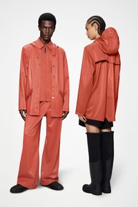 Αδιάβροχο κοραλλί μπουφάν βροχής και παντελόνια με λαμπερό φινίρισμα, συνδυασμένα με μαύρες γαλότσες μέχρι το γόνατο, παρουσιασμένα σε δύο μοντέλα.