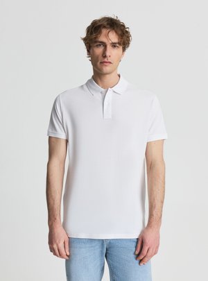 Giovane uomo con capelli ricci che indossa una polo bianca semplice a maniche corte e jeans azzurri su uno sfondo neutro.
