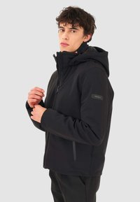 Giacca nera con cappuccio realizzata in materiale morbido e elastico. Presenta una chiusura lampo frontale, tasche laterali e una toppa con logo sul braccio sinistro.