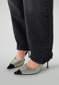 Chaussures à talons hauts argentées scintillantes avec un bout en cuir verni noir. Portées avec un pantalon en denim noir retroussé.