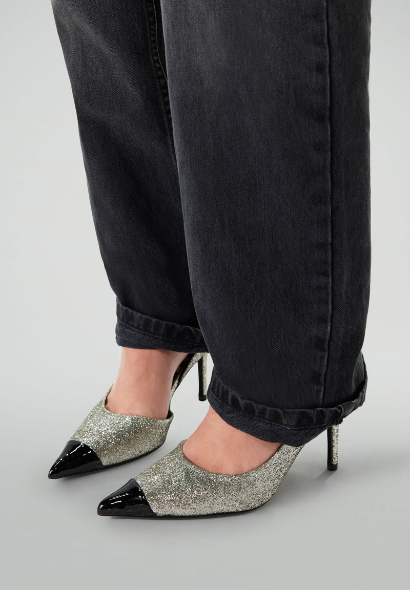 Chaussures à talons hauts argentées scintillantes avec un bout en cuir verni noir. Portées avec un pantalon en denim noir retroussé.