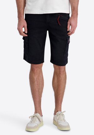 Mann trägt schwarze Cargoshorts und weiße Sneakers, steht mit entspannten Händen an den Seiten vor einem einfarbigen Hintergrund.