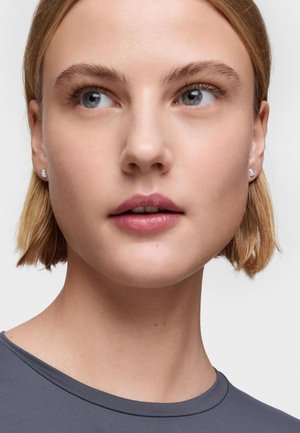 Primo piano di una persona con i capelli corti e biondi, che indossa una maglietta grigia. Caratteristiche degne di nota includono chiari occhi azzurri e orecchini a perno discreti.