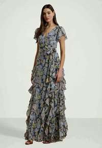 Mujer vistiendo un vestido floral de manga corta y largo hasta el suelo, con volantes y un lazo lateral, de pie frente a un fondo claro y liso.