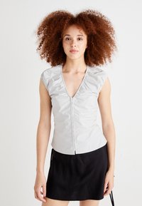 Top argent sans manches avec un col en V et des détails froncés aux épaules. Texture lisse, silhouette ajustée, associé à une jupe noire.