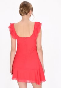 Robe rouge avec des manches volantées, créant une texture en couches. Présente un dos nu, une fermeture éclair et un ourlet évasé au-dessus du genou.