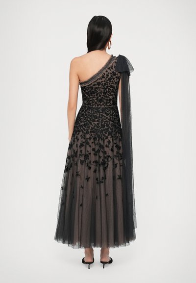 Femeie cu spatele, îmbrăcată într-o rochie de seară neagră cu un umăr, având un overlay din dantelă florală și o eșarfă transparentă drapată pe umăr.