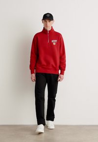 Pulover roșu cu guler înalt și fermoar, având un logo, asortat cu pantaloni negri și adidași albi. Design minimalist.