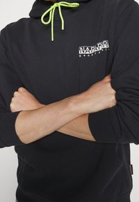 Sudadera negra con capucha y logo frontal, con cordón verde moteado, puños acanalados y bolsillo tipo canguro. Textura de tela suave.