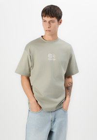 Ung mand med kort brunt hår iført en lys grøn oversized t-shirt med logo og lyseblå jeans, hænderne i lommerne, stående mod en hvid baggrund.