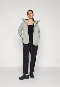 Giacca a vento verde chiaro con cappuccio, canotta nera, pantaloni cargo neri e sneakers nere. La giacca presenta una vestibilità rilassata e tasche con zip.