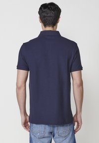 Jūros jėgos polo marškinėliai su trumpomis rankovėmis ir raukinėtu apykakle, turintys lygią tekstūrą. Marškiniai turi tiesią apatinę dalį ir derinama su šviesiais džinsais.