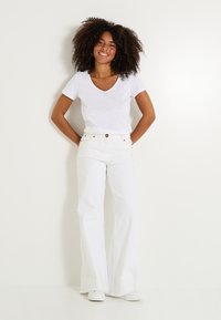 Camiseta blanca ajustada con escote en V combinada con pantalones vaqueros acampanados blancos de cintura alta. La modelo está de pie con las manos en las caderas, usando zapatillas blancas.