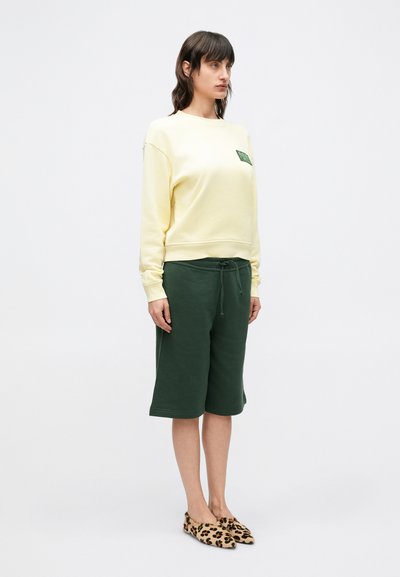 Κίτρινο φούτερ με ένα μικρό πράσινο μπαλώμα, συνδυασμένο με σ κούρα πράσινη βερμούδα και παπούτσια τύπου slip-on με λεοπάρ μοτίβο, με άνετη εφαρμογή.