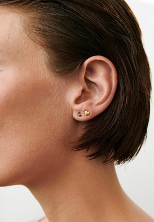 Close-up af en persons øre med kort brunt hår, iført to små guldørestikker, én hjerteformet og én halvmåneformet med en ædelsten.
