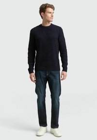 Dunkelblauer Strickpullover mit gesprenkelter Textur, Rundhalsausschnitt, kombiniert mit dunklen Jeans und weißen Sneakers, steht vor einem neutralen Hintergrund.