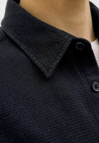 Chemise noire à boutons en tissu texturé, semblable à un gaufre. Dotée d'un col structuré et de deux boutons noirs à l'avant.
