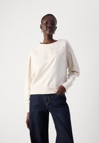 Felpa color crema con un motivo floreale testurizzato sulla parte superiore del petto, maniche ampie e orlo a costine, abbinata a pantaloni in denim scuri a gamba larga.