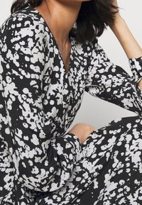 Musta mekko valkoisilla abstrakteilla täplillä, V-pääntiellä, pitkillä hihoilla ja väljä istuvuus. Kangas vaikuttaa pehmeältä ja sileäpintaiselta.