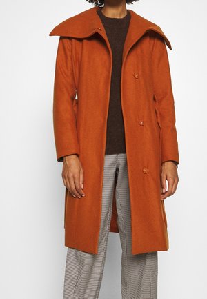Oranžový vlněný kabát s širokým límcem, předními knoflíky a postranními kapsami, doplněný hnědým svetrem a kostkovanými kalhotami.