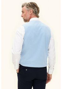 Hellblauer Wollsweater-Weste, die über einem weißen Hemd getragen wird. Sie hat ein klassisches Design und eine glatte Textur, dazu sind marinefarbene Hosen sichtbar.