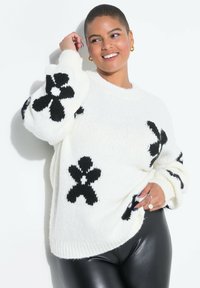 Weißer Strickpullover mit schwarzen Blumenmustern, mit gerippten Bündchen und Saum. Kombiniert mit eleganten schwarzen Leggings. Strukturierter, gemütlicher Stoff.