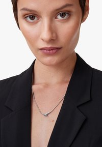 Silver necklace with a pendant resembling a bird, worn over a black blazer. Smooth metal chain and a sleek, contemporary design.