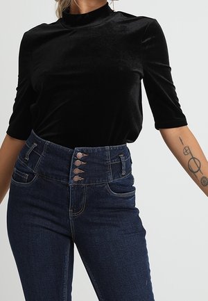 Kobieta w czarnej aksamitnej bluzce z krótkim rękawem i ciemnoniebieskich dżinsach z wysokim stanem na cztery guziki, z widocznym tatuażem na lewym przedramieniu.