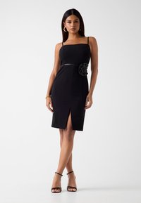 Schwarzes Kleid mit quadratischem Ausschnitt, taillierter Passform, seitlichem Schlitz und einem Stoffblumenakzent an der Taille. Glatte Textur und verstellbare Träger.