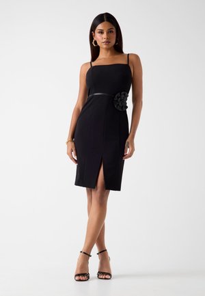 Zwarte jurk met een vierkante halslijn, getailleerd ontwerp, zijsplit en een stoffen bloemaccent op de taille. Soepele textuur en verstelbare bandjes.