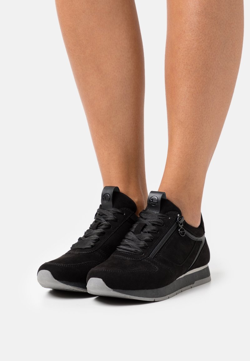 Baskets en daim noir avec lacets noirs, fermeture éclair latérale et semelles grises portées sur des jambes nues sur fond blanc.