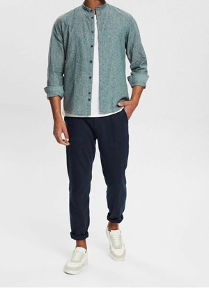 Man wearing een groen overhemd met patroon over een wit T-shirt, donkerblauwe opgerolde broek en witte sneakers tegen een effen achtergrond.