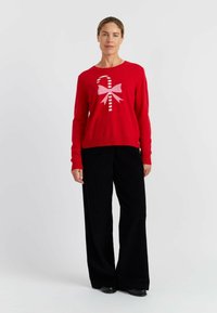Pull en tricot rouge avec un nœud rose et un motif de canne en sucre sur le devant, associé à un pantalon en velours côtelé noir à coupe large et des chaussures noires.