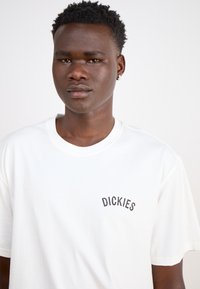 White cotton t-shirt featuring a round neckline and short sleeves. Displays the word "DICKIES" in black arch font on the left chest.