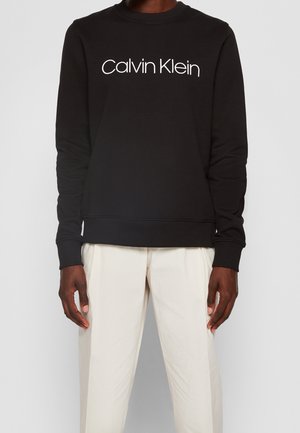 Sweat-shirt noir à manches longues avec le logo blanc Calvin Klein sur la poitrine, porté avec un pantalon beige clair par une personne debout devant un fond uni.