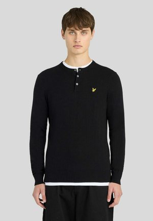 Jeune homme portant un t-shirt henley noir à manches longues avec trois boutons et un petit logo d’oiseau jaune sur la poitrine.