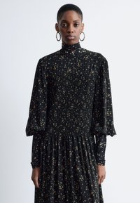 Schwarzes Kleid mit floralem Muster, hohem Kragen, gerafftem Oberteil und voluminösen Ärmeln. Strukturiert mit dezenten Falten im gesamten Stoff.