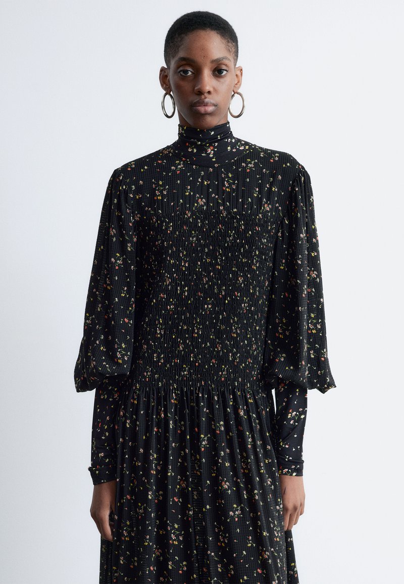 Schwarzes Kleid mit floralem Muster, hohem Kragen, gerafftem Oberteil und voluminösen Ärmeln. Strukturiert mit dezenten Falten im gesamten Stoff.