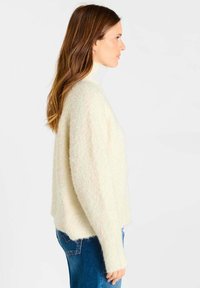 Vrouw met lang bruin haar, gekleed in een witte pluizige coltrui en blauwe jeans, staand in profiel tegen een effen achtergrond.