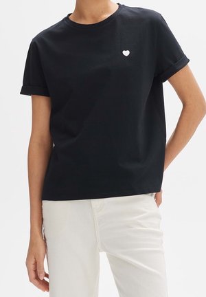 Frau trägt ein schwarzes kurzärmeliges T-Shirt mit einem kleinen weißen Herz auf der linken Brust, kombiniert mit weißer Hose.