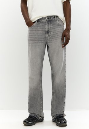 Mann trägt hellgraue, gerade geschnittene Jeans, schwarze Sneakers und ein weißes Hemd, steht mit den Händen in den Taschen vor einem schlichten Hintergrund.