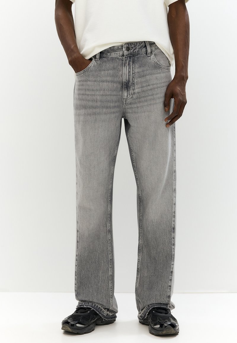 Homme portant un jean droit gris clair, des baskets noires et une chemise blanche, les mains dans les poches, debout devant un fond uni.