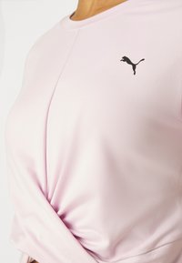 Top deportivo rosa claro con cuello redondo, diseño elegante, detalle de nudo en la cintura y logo de Puma negro en el lado izquierdo del pecho. Textura de tela suave.