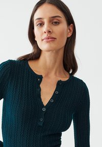 Teal ribbed long-sleeve top with a henley neckline, featuring four dark buttons and a textured pattern across the fabric.