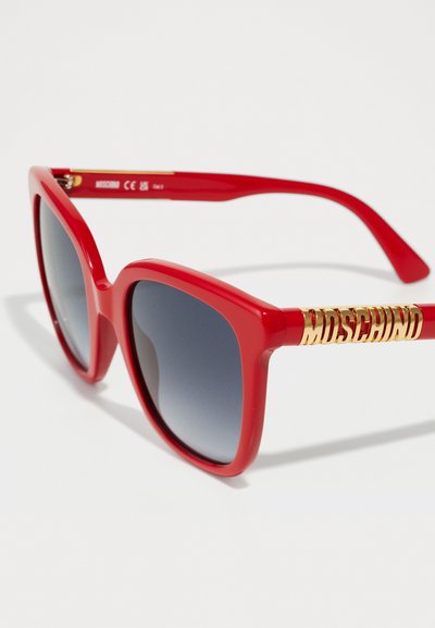 Ochelari de soare din plastic roșu, cu rame groase și pătrate și lentile gri cu gradient. Prezintă un logo auriu "Moschino" pe tâmplă.