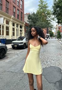 Yellow mini dress with thin straps, smooth texture, and fitted bodice. Background features brick buildings and a cobblestone street.